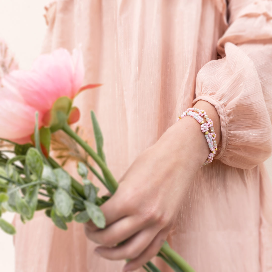 Bild von einem Model in einem rosa Kleid. Im Vordergrund ist eine Hand mit zwei DIY Armband mit Blumen Armbändern in pastellfarben. Die Perlen der Armbänder sind in Form von Blüten angeordnet.